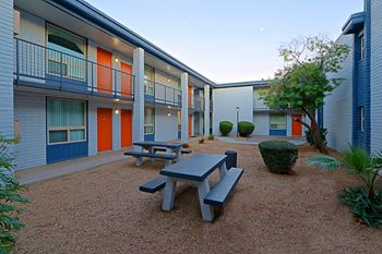Picnic area with woodchip ground, two grey picnic tables, surrounded yb apartments with red doors and a blue balcony railing at Allora Phoenix in Phoenix, Arizona